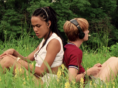 Two young women sitting in a field, sitting back to back.