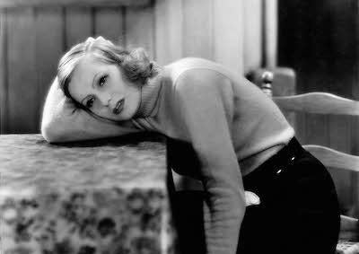 Greta Garbo looking distraught, resting her arm and head on a table.