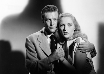 A man embracing a frightened woman, a shadow looming behind them.