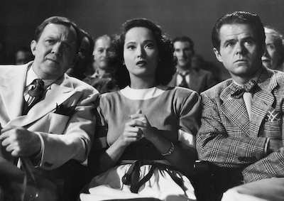 Merle Oberon sitting among a crowd.