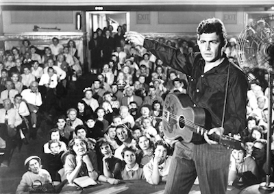 A man holding a guitar in front of a seated audience. 