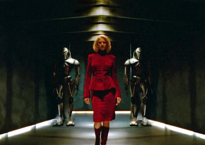 A woman in a red dress walking in front of two figures in armor.