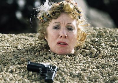 A woman buried up to her neck in the ground, with a gun near her head.