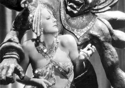Greta Garbo in a glittering costume and headdress in front of a multi-armed statue.