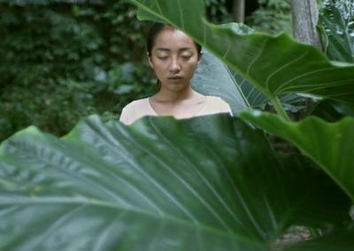 A person standing with eyes closed behind large leaves.