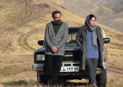 A man and a woman leaning against a car in an empty, hilly landscape.