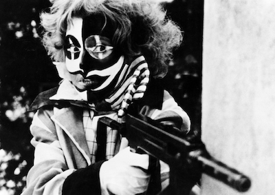 A person in harlequin style makeup and holding a gun.