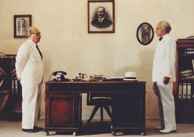 Two men standing and facing each other from across a desk.