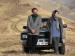 A man and a woman leaning against a car in an empty, hilly landscape.