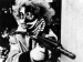 A person in harlequin style makeup and holding a gun.